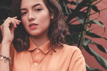 an image of a young person with brown hair to their shoulders wearing a pinkish orange shirt